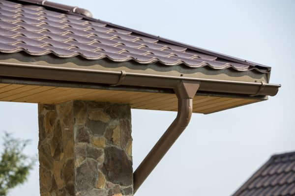 Detail of new modern house cottage corner with stucco walls decorated with natural stones, brown shingled roof and rain gutter pipe system on blue sky copy space background. Real estate concept.